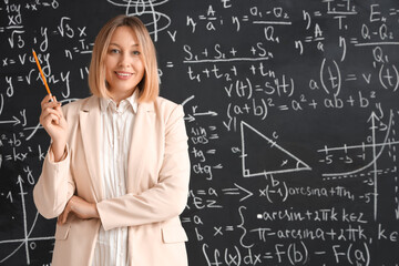 Mature female math teacher with pencil near blackboard in classroom
