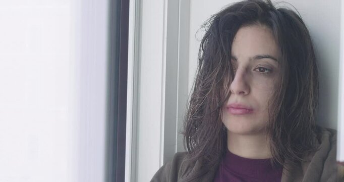 A close-up of a young girl at sunrise after a bad night, tears in her eyes and smeared makeup, standing by the window, feeling very sad.