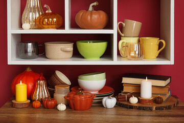 Wooden table with different dishes, books, autumn decor and stylish shelf hanging on color wall