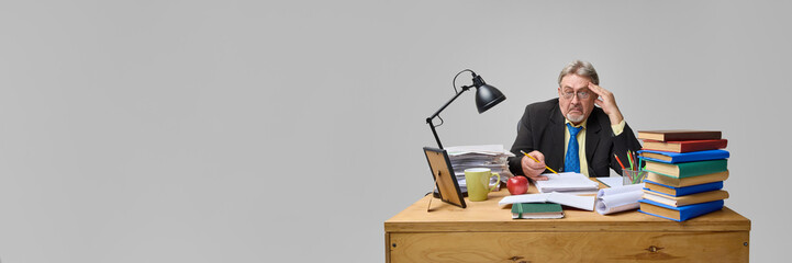 Shocked senior man, teacher sitting at messy desk and looking at papers with confusion against light grey background. Checking students works. Concept of education, profession, learning