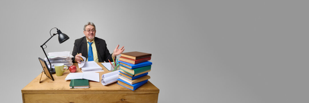 Senior businessman gesturing expressively at overloaded desk with documents and books on grey background. Concept of corporate stress, education, financial reports, office chaos, profession