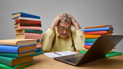 Tired elderly teacher with hands on head at cluttered desk with laptop and books against grey...