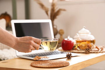 Female hand holding cup of tea near laptop on table in room. Closeup