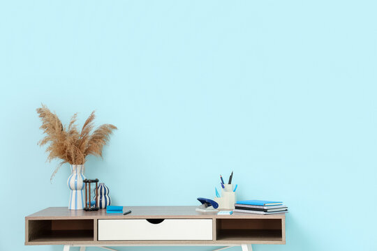 Stylish workplace with vase of pampas grass, stationery and hourglass near blue wall in comfortable office