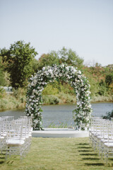 A beautiful outdoor wedding ceremony setup by the river with a floral arch and transparent chairs. The elegant white and green decor blends with the natural landscape