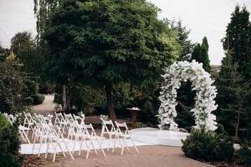 A small courtyard surrounded by trees, featuring a white flower and greenery arch, a white pathway,...