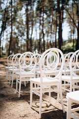 Rows of white modern chairs with round backrests stand on a sandy ground amidst tall trees in an...