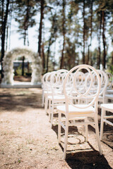 Rows of white modern chairs with round backrests stand on a sandy ground amidst tall trees in an...