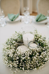 Thick white candles in glass candle holders and white-green floral compositions decorating a banquet table outdoors, creating an elegant and serene atmosphere for the celebration