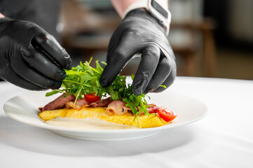 A chef wearing black gloves delicately garnishes a delicious omelette on a white plate, adding...