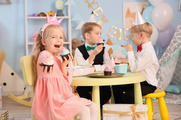 Cute little happy girl and her friends with cupcakes celebrating birthday at table in children's room