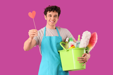 Happy male janitor with bucket of cleaning supplies and paper heart on purple background