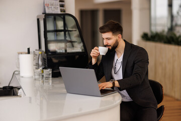 Confident young man working on laptop and holding coffee cup while sitting at his working place in office