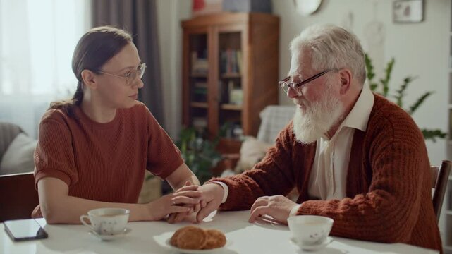 Young woman sitting at table with teacups, holding hand of elderly grandfather during heartfelt conversation, providing care and emotional support at home