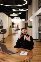 Happy manmanager working remotely talking on mobile phone, sitting at desk in office interior