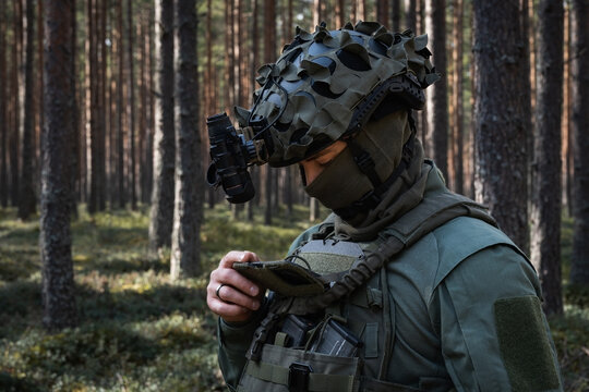 A soldier in a tactical helmet with a night vision device and balaclava uses a mobile phone, attached to his gear, for navigation in the forest.