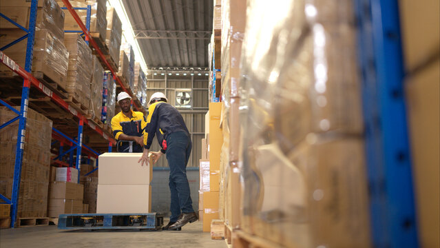 Two warehouse workers are lifting boxes of merchandise on a pallet jack to store on a shelf in a warehouse