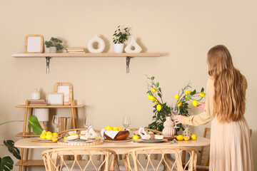 Young woman decorating table setting with fresh citruses, lemon tree branches and glasses in dining room, back view