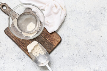 Bowl with flour, metal sieve, scoop and wooden board on light background