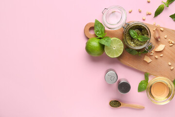 Wooden board with jar of delicious pesto sauce and ingredients on pink background