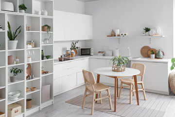 Interior of stylish kitchen with white counters, table, houseplants and shelving unit