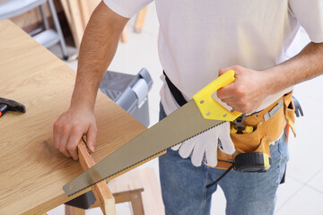 Male construction worker with professional tools and handsaw sawing wooden plank at table in workshop, closeup