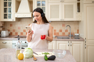 Woman holding a tomato and looking at her phone in the kitchen. She wears headphones, multitasking...