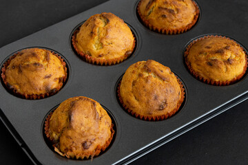 Freshly baked muffins in a muffin baking pan on dark background. Warm cupcakes in paper cups in muffin tin, baked homemade treats having a well-risen texture and slight crustiness