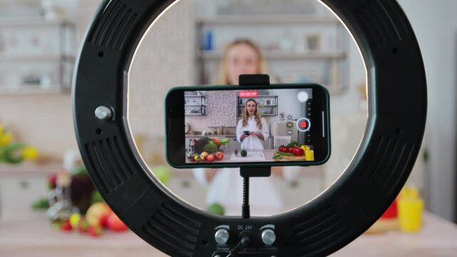 Young woman food blogger cooking salad in front of smartphone camera while recording vlog video and live streaming at home in kitchen.