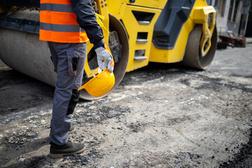 Construction worker stands next to road roller while holding safety helmet on job site