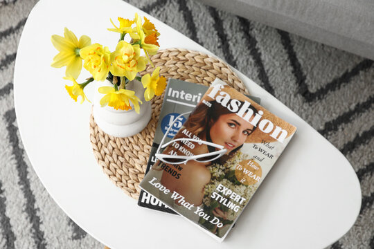 Vase of beautiful daffodil flowers with sunglasses and magazines on coffee table in living room, top view