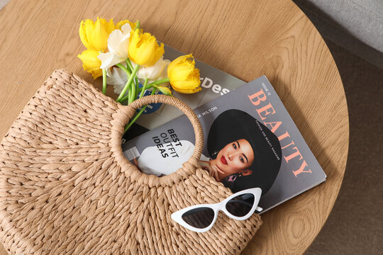 Wicker bag of beautiful tulip flowers with magazines and sunglasses on coffee table in living room, top view
