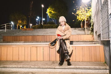 A woman in a white coat and beanie using a smartphone on city steps at night, ideal for urban,...