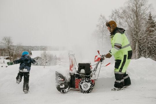 Side view of man using snowblower while son playing during winter