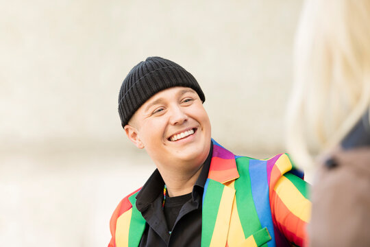 Happy gay person wearing multi colored jacket and knit hat talking with friend