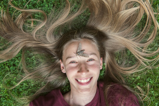 Directly above portrait of happy girl lying on grass with hawk moth on face