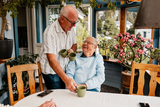 Senior man giving mug to male friend sitting at table in patio