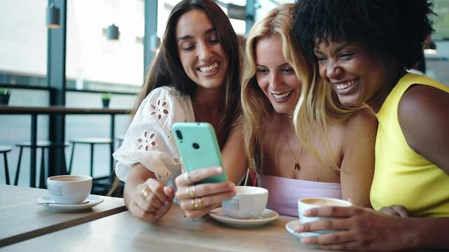 Three cheerful female gil friends looking at a mobile phone app and laughing while sitting in a cafe