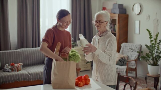 Young woman assisting elderly grandmother with everyday routine, unpacking groceries on table, smiling and chatting at home