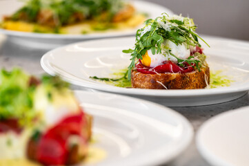 A gourmet dish with a slice of toasted bread topped with a poached egg, fresh greens, red vegetables, and grated cheese, served on a white plate.