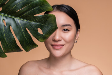 Beautiful young woman girl model with clean fresh skin touching her face in tropical green leaf looking at camera posing standing isolated beige background . Cosmetology beauty and spa care concept.