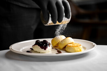 A chef in black gloves carefully sprinkles powdered sugar over three delectable cheesecakes served with a blueberry sauce on a white plate in a stylish kitchen.