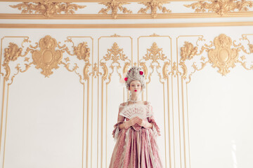 Elegant woman in vintage gown with intricate hairstyle and fan stands in ornate palace interior, blending historic and contemporary styles