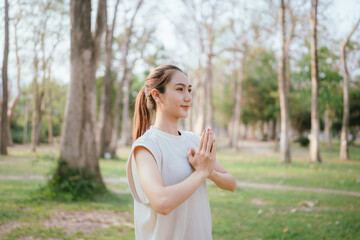 Fototapeta premium Young Asian woman practicing yoga in a quiet park, gracefully in a relaxed pose, relaxing and strengthening her body, flexibility and relaxation amidst the green grass.