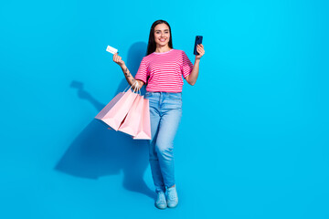 Smiling young woman holds shopping bags, a credit card, and her phone against a vibrant blue...