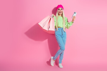 Fashionable young woman with shopping bags and phone posing against pink background wearing trendy attire and beanie