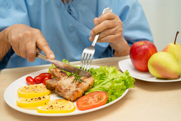 Asian senior woman patient eating pork chop stake and vegetable salad for healthy food in hospital.