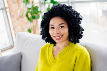Young woman with curly hair wearing a bright green jumper on a comfortable sofa, with a cheerful...