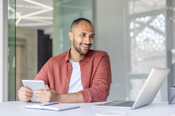 A smiling man sits at a desk, using a laptop and holding a tablet. Natural light floods the modern...