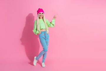 Fashionable young woman in vibrant green pullover and striped pants posing against a pink background, embracing casual style
